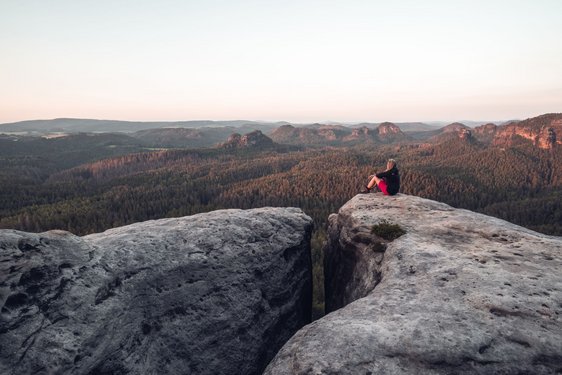 Eine Frau sitzt auf einem Felsen in der Sächsischen Schweiz und blickt über die umliegenden Wälder.