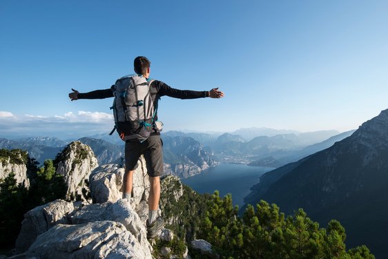 Ein Wanderer steht mit weit ausgebreiteten Armen im Gebirge über dem Gardasee und schaut hinunter.