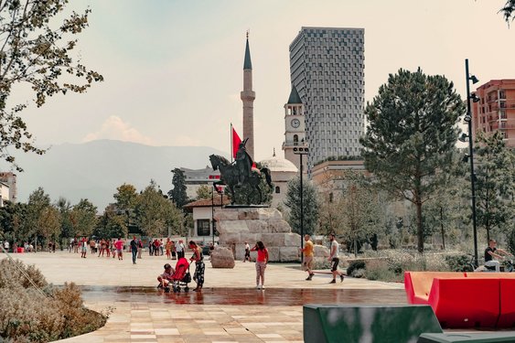 Im Vordergrund des Platzes sind viele Menschen unterwegs, im Mittelgrund steht ein Reiterstandbild von Skanderbeg, im Hintergrund sieht man eine Moschee mit einem Minarett und ein Hochhaus.