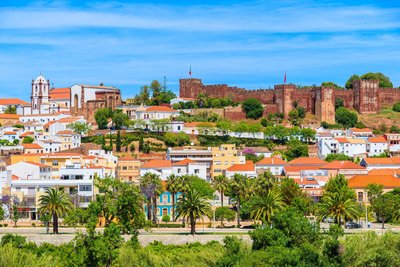 Blick über das Zentrum von Silves Man sieht Häuser, die sich einen Hügel hinaufziehen, der von einer Burg gekrönt wird; im Vordergrund eine von viel Grün flankierte Straße.