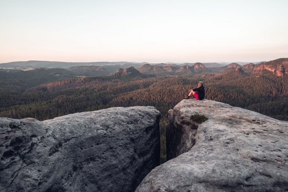 Weiter Ausblick über die Sächsische Schweiz Eine Frau sitzt auf einem Felsen in der Sächsischen Schweiz und blickt über die umliegenden Wälder.
