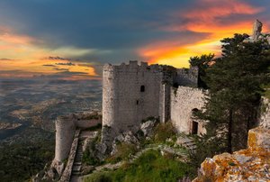 Die Festung Kantara auf Zypern Auf einer Anhöhe liegt eine Festung, hinter ihr schaut man hinab über eine weite Ebene; der Himmel ist vom Sonnenuntergang in orange getaucht, darüber graue Wolken.