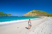 Man sieht eine sommerlich gekleidete Frau vom Betrachter weg über einen Sandstrand in einer Bucht auf Lombok spazierengehen. 