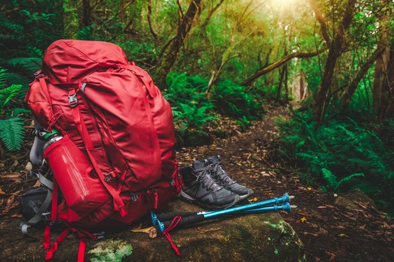 Auf dem Waldboden steht ein roter Wanderrucksack nebst -schuhen und -stöcken.