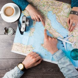 Zwei Menschen, von denen man nur die Hände sieht, sitzen über einer Landkarte von Italien, daneben Utensilien, die auf Planungen verweisen wie ein Miniaturflieger und ein Kompass.