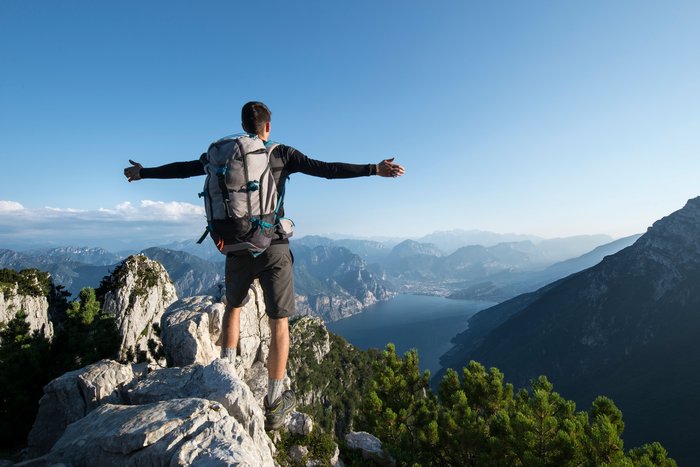 Ein Wanderer steht mit weit ausgebreiteten Armen im Gebirge über dem Gardasee und schaut hinunter.