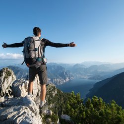 Ein Wanderer steht mit weit ausgebreiteten Armen im Gebirge über dem Gardasee und schaut hinunter.
