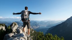 Ein Wanderer steht mit weit ausgebreiteten Armen im Gebirge über dem Gardasee und schaut hinunter.