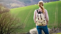 Man sieht die Rückensicht einer Frau it Wanderrucksack, die vor einem Feld steht.