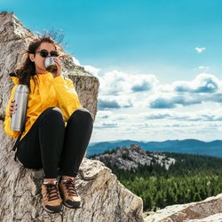 Eine Frau in Wanderkleidung sitzt auf einem kargen Berggipfel und trinkt etwas aus einer Thermoskanne.