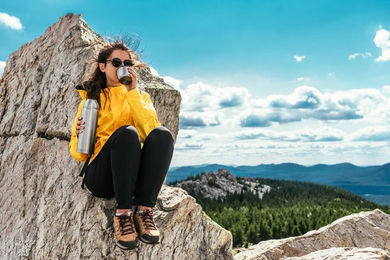 Eine Frau in Wanderkleidung sitzt auf einem kargen Berggipfel und trinkt etwas aus einer Thermoskanne.