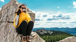 Eine Frau in Wanderkleidung sitzt auf einem kargen Berggipfel und trinkt etwas aus einer Thermoskanne.