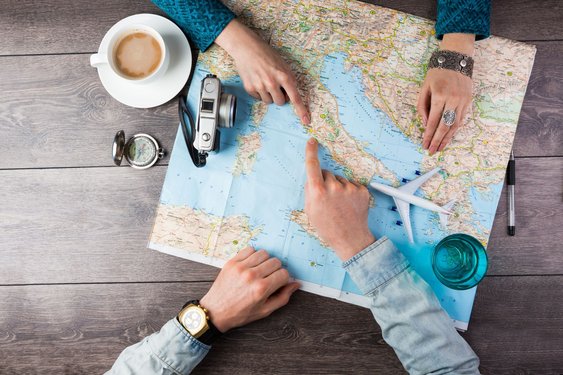 Zwei Händepaare auf einer Europakarte zeigen auf verschiedene Regionen. Außerdem im Bild: eine Kamera, ein Kompass, ein Modellflugzeug und Getränke.