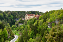 In der Bildmitte liegt eine Burg auf einer Anhöhe umgeben von Wäldern. Das obere Drittel des Bildes nimmt der Himmel ein.
