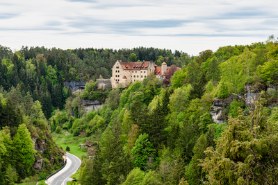 Umgeben von den Wäldern der Fränkischen Schweiz: Burg Rabenstein In der Bildmitte liegt eine Burg auf einer Anhöhe umgeben von Wäldern. Das obere Drittel des Bildes nimmt der Himmel ein.