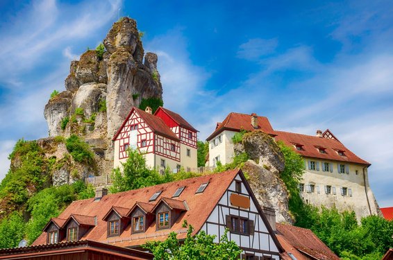 Zwei Fachwerkhäuser und ein burgartiges Gebäude gruppieren sich unter einer auffälligen Felsformation.