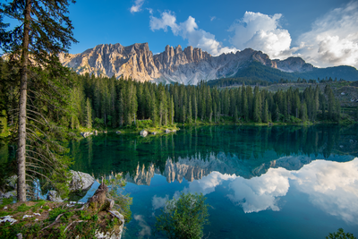 Das Latemar-Massiv oberhalb des Karersees in Südtirol In einem von Bäumen umstandenen Gebirgssee spiegelt sich ein Gebirgsmassiv sowie der darüber befindliche blaue Himmel mit weißen Wolken rechts.