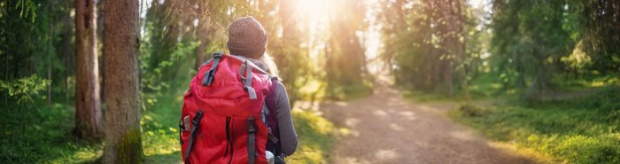 Eine Frau mit einem großen roten Rucksack ist wandern in einem Tannenwald unterwegs, der von Sonne durchschienen ist.
