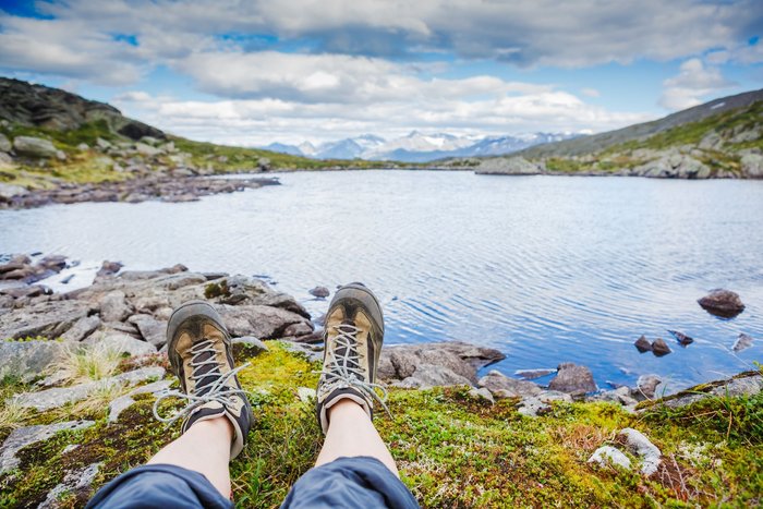 Vor einem Seepanorama sieht man nur die mit Wanderschuhen bekleideten Unterschenkel am Ufer ausgestreckt.