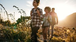 Drei Wanderer laufen über einen Feldweg während im Hintergrund die Sonne untergeht.