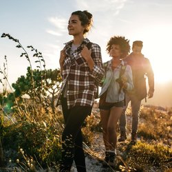 Drei Personen laufen im Vordergrund durch ein Feld, während im Hintergrund die Sonne hinter einem Berg verschwindet.
