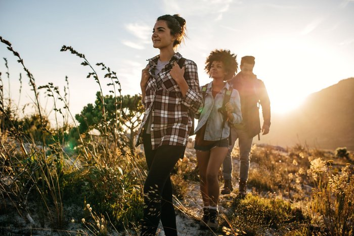 Drei Personen laufen im Vordergrund durch ein Feld, während im Hintergrund die Sonne hinter einem Berg verschwindet.