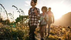 Drei Personen laufen im Vordergrund durch ein Feld, während im Hintergrund die Sonne hinter einem Berg verschwindet.