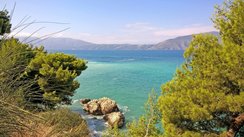Zwischen Büschen hindurch schaut man auf das türkisblaue Meer der Adria; im Hintergrund erheben sich sanfte Berge.