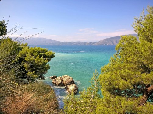 Zwischen Büschen hindurch schaut man auf das türkisblaue Meer der Adria; im Hintergrund erheben sich sanfte Berge.
