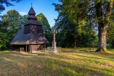 In der linken Bildhälfte steht eine kleine aus Holz gebaute Kirche auf einer Wiese. Die UNESCO-Welterbestätte ist von Bäumen umgeben.