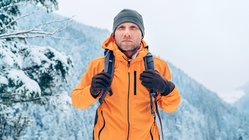 Ein Mann in einer orangenen Softshelljacke schreitet durch eine verschneite Berglandschaft.