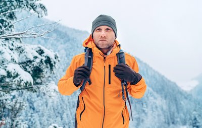 Ein Mann in einer orangenen Softshelljacke schreitet durch eine verschneite Berglandschaft.