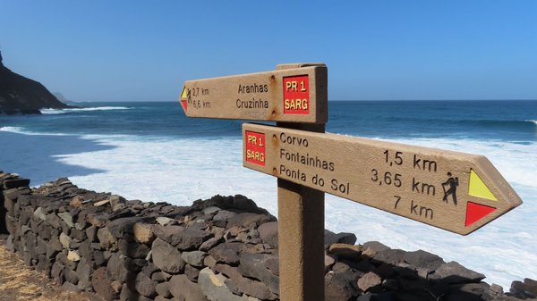 Wandermarkierungen auf Santo Antão Vor dem weiß-blauen Meer steht ein Wanderwegweise, der die Entfernungen zu verschiedenen Orten auf Santo Antão angibt.